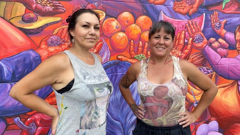 Two women pose side by side in front of a warm-coloured mural they painted. The section of the mural captures shows women holding out their hand carrying different seats over some fruits and vegetables. 