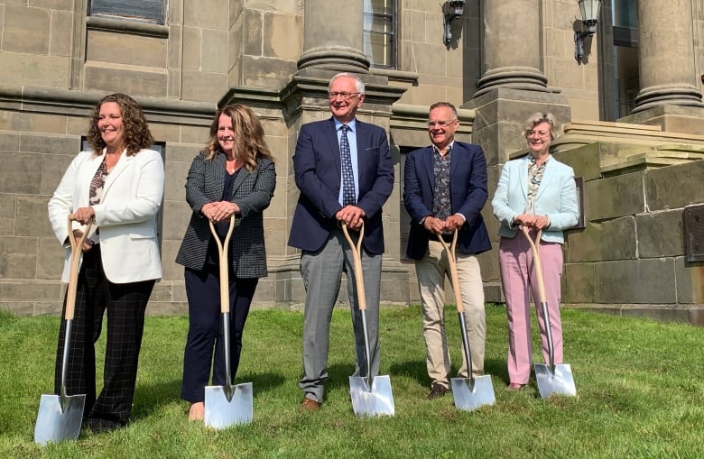 Five individuals, three women, two men, standing side by side with shovels in the grass smiling at camera. 