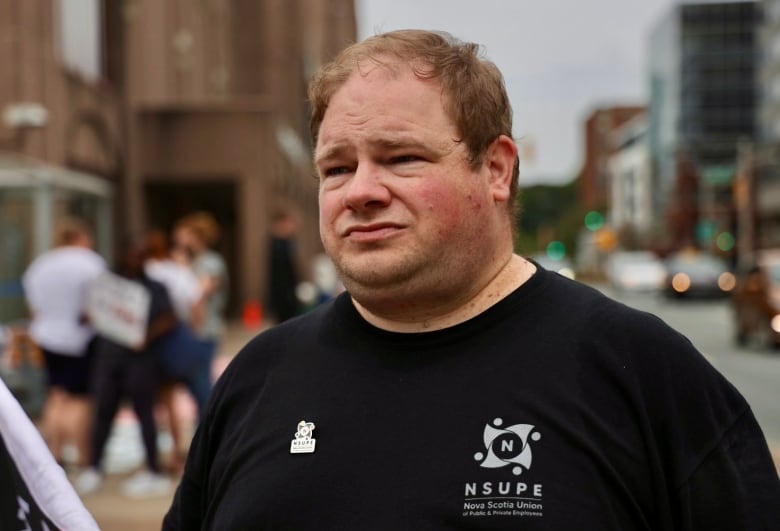 man in black t-shirt stands near people holding signs on a sidewalk