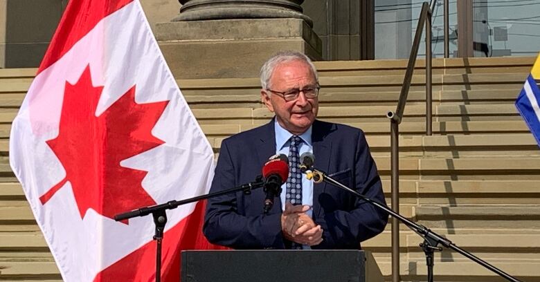 Older man wearing suit and tie with neutral expression standing with building behind him on a sunny day. 