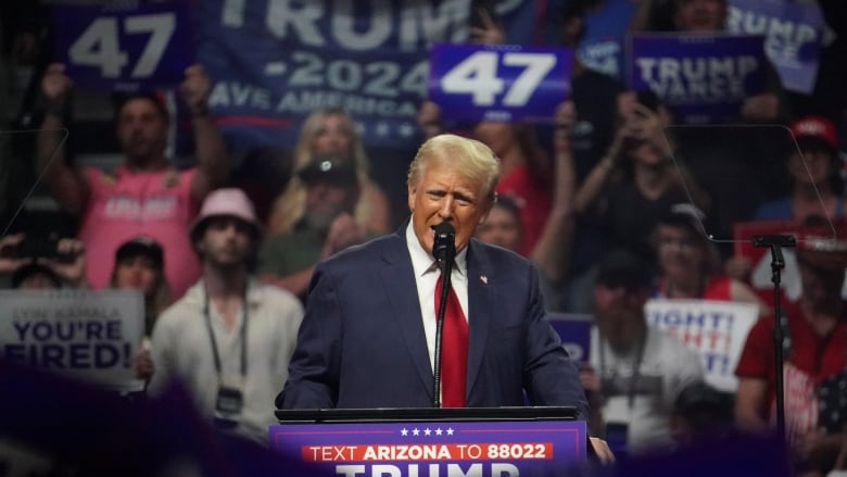 A man in a dark suit a long red tie stands at a podium.