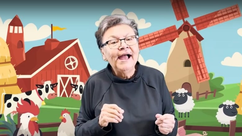 A woman in a black shirt stands in front of a cartoon farm scene depicting a barn, windmill, fields, sheep, cows, chickens, and a pig. 