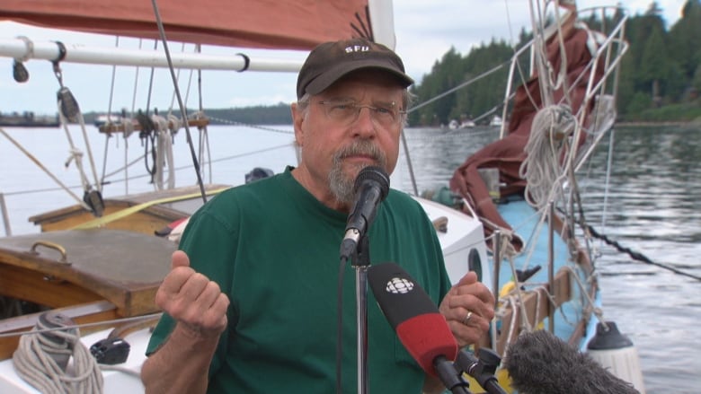 A man wearing a green shirt gestures as he speaks in front of a ship moored on the water.