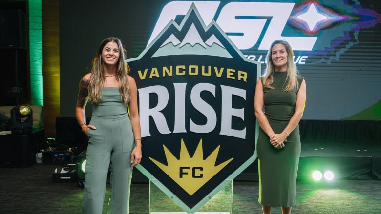 Two white woman smile and pose next to a teal symbol of three mountains, and the words 'Vancouver Rise FC'.