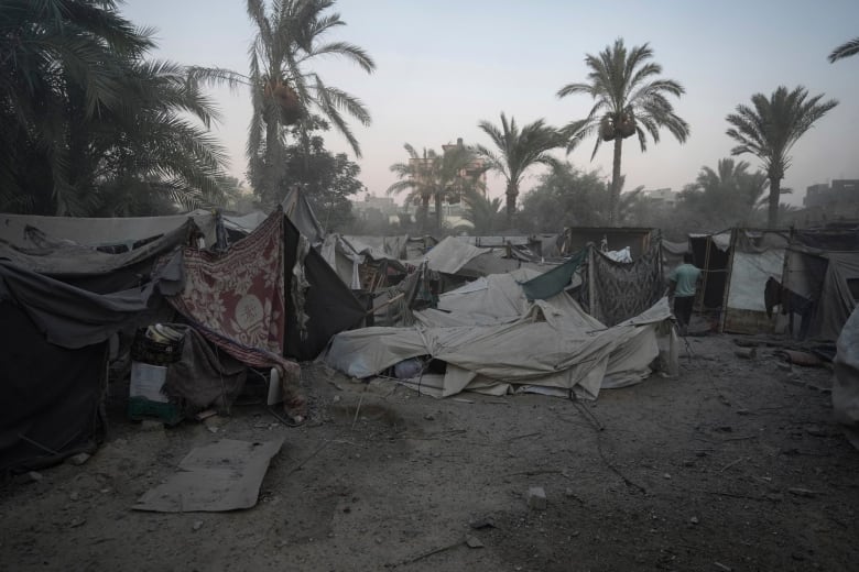 Tents covered in grey dust and rubble are pictured. A row of trees lie beyond the circle of ruined tents. 