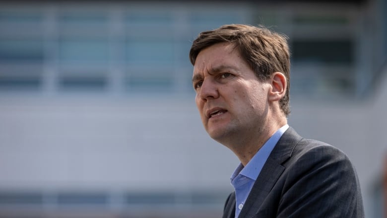 B.C.'s Premier David Eby wearing a dark grey suit standing behind a white, tall building. 