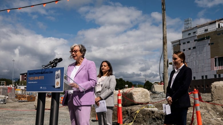 Three women standing near a podium outside in front of a hospital. 