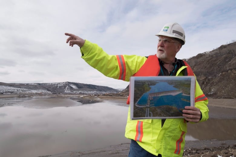 A man wearing a high-vis vest points to his right while a snowy water body is seen behind him.