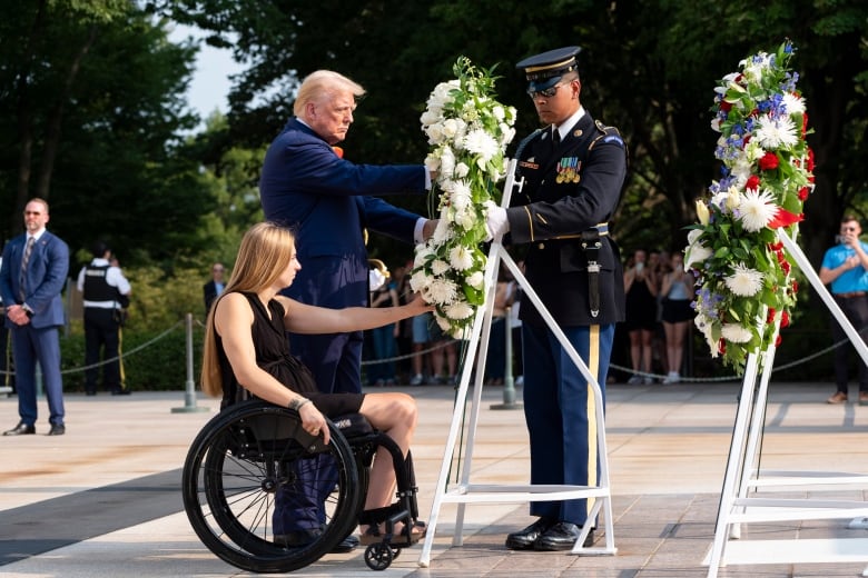 A woman in a wheelchair and man in a blue suit lay a wreath with the help of a man in uniform.