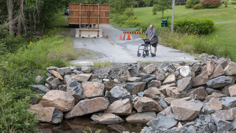 A man with a walker stands in the middle of a road looking down at a large gap where there was once a bridge. The gap is lined with rocks and a stream running down the middle.