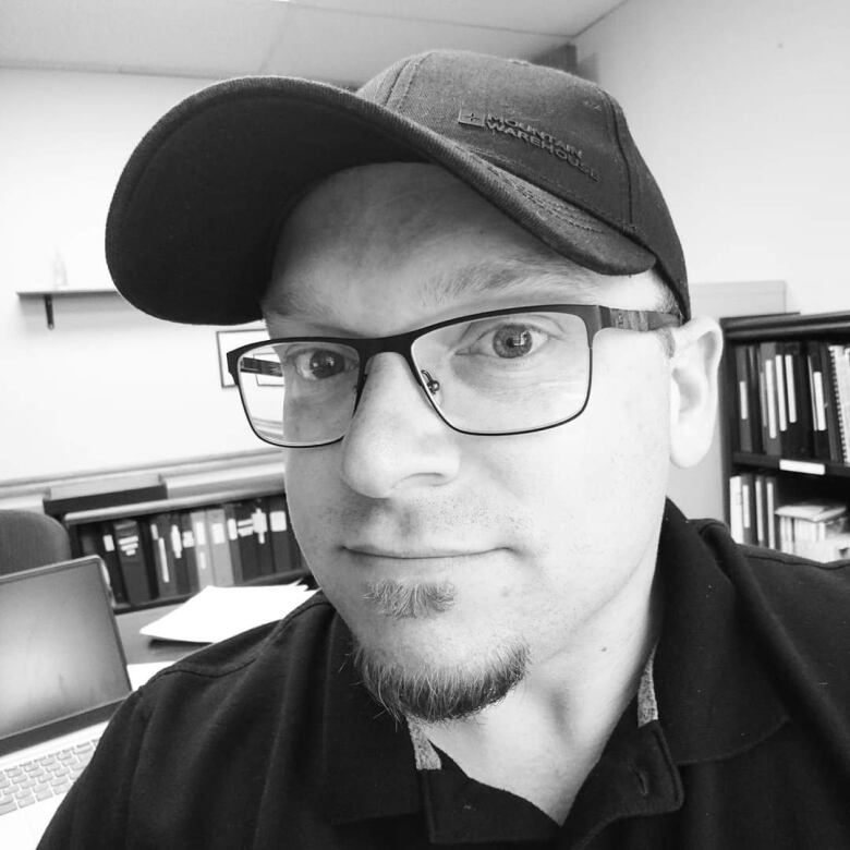 A black and white photo of a man's face. He has a goatee, glasses and is wearing a baseball cap. There are files on shelves behind him.