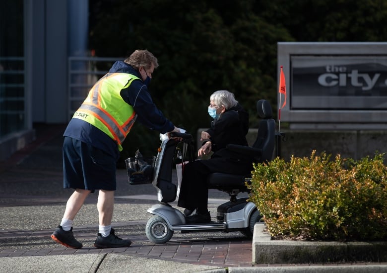 A man in a yellow vest helps an elderly woman on a mobility scooter.