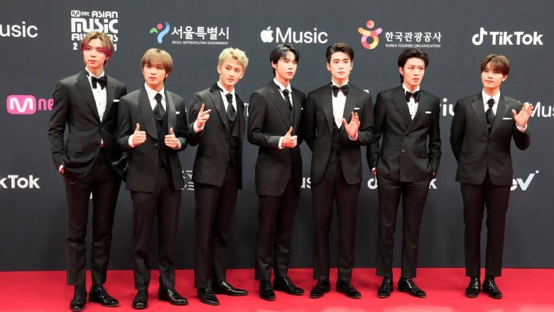 A group of korean boyband is standing in a line wearing suits facing the camera. 