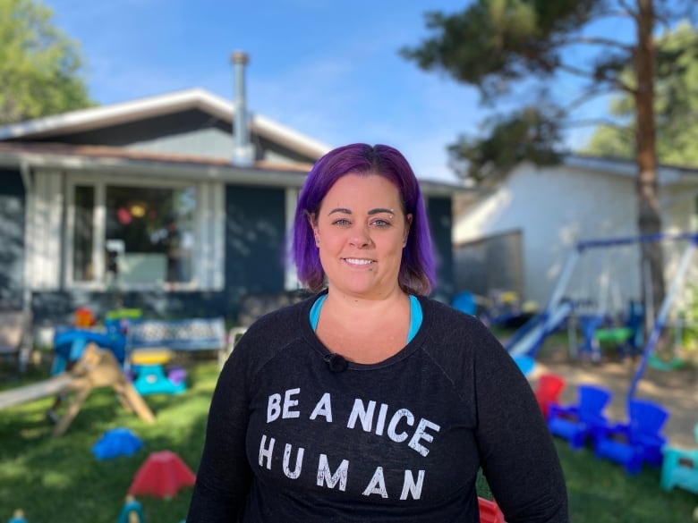 A woman  with purple colored hair and a dark shirt stands outside in front of a yard full of daycare outside toys.