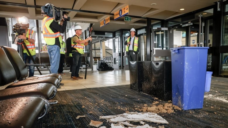 Debris from a broken ceiling is pictured on the floor of an airport with a man wearing a high visibility vest and construction helmet pictured in the foreground and background.