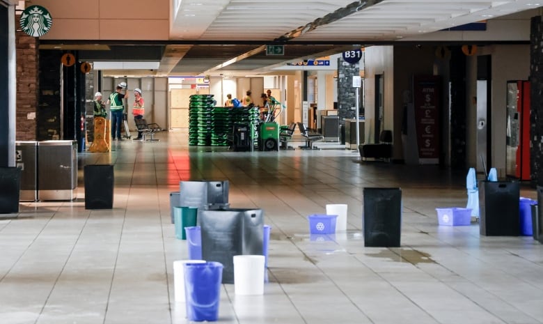 Blue, white and black buckets collecting rainwater are place on the floor of an airport with construction workers pictured in the background.