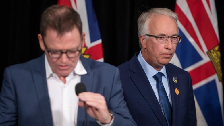 Two men speak with B.C. flags in the background. One of them is in focus and looks pensive, while the other looks down at the mic and forlorn.