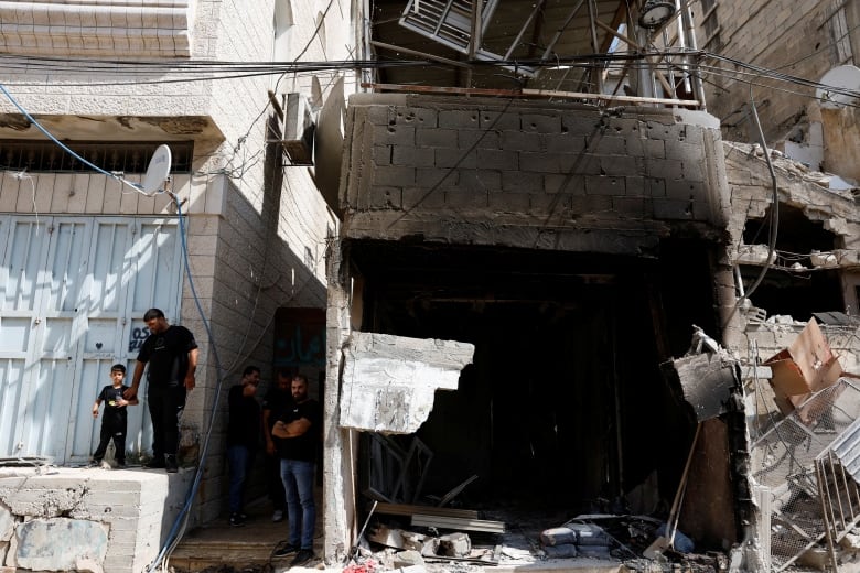 A man holds the hand of a small child next to a blackened structure with a massive hole in the front of it. 