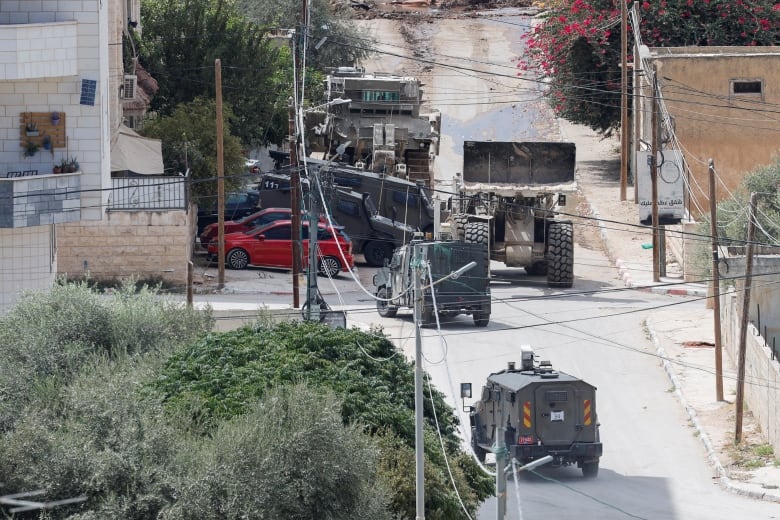 Several different types of military vehicle move down a street. 