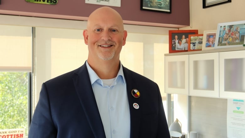 A bald man in a dark blue jacket and light blue dress shirt smiling in an office with various photos in the background.