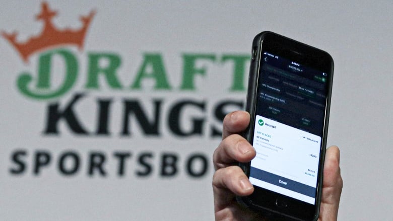 A phone is pictured being held up by a hand in front of a sign that reads DraftKings.