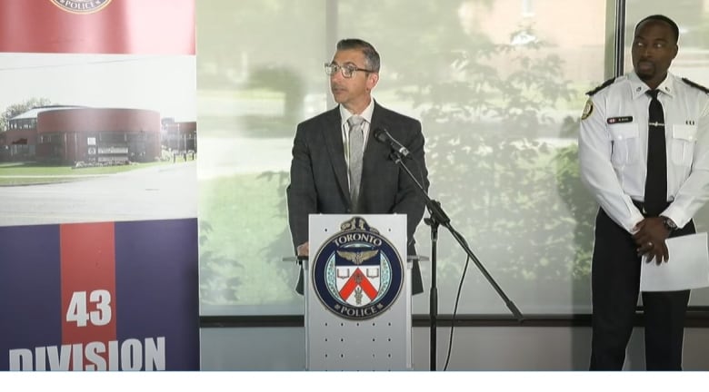 A man in a suit stands at a Toronto police podium indoors, next to a uniformed police officer