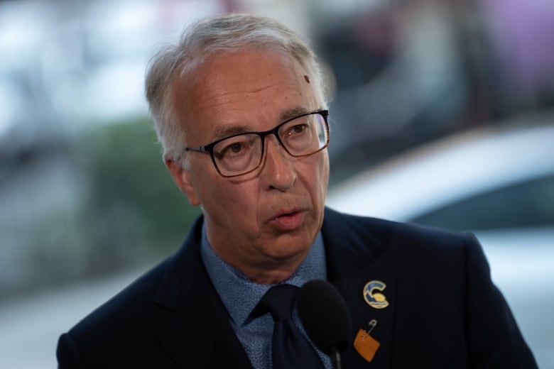 A white-haired man in glasses wearing a dark suit jacket over a dark blue shirt.