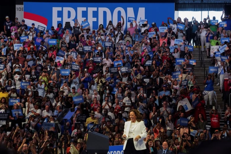 Harris in white suit, speaking in front of crowd with blue signs