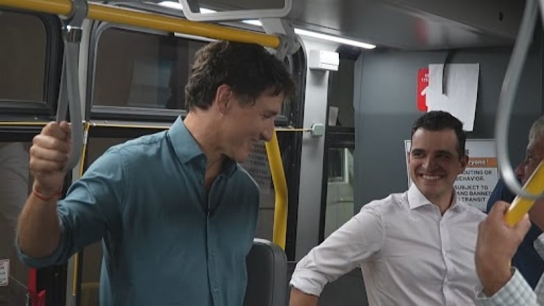 A male politician in a blue shirt stands on a transit bus talking to another politician in a white shirt.