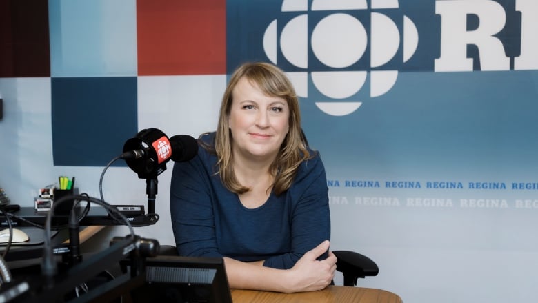 A brunette Caucasian woman in a blue tee shirt leans on a news desk behind a mic inside a radio studio