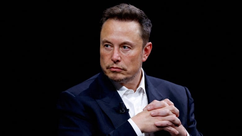 A man sits with his hands folded in front of a black backdrop. 