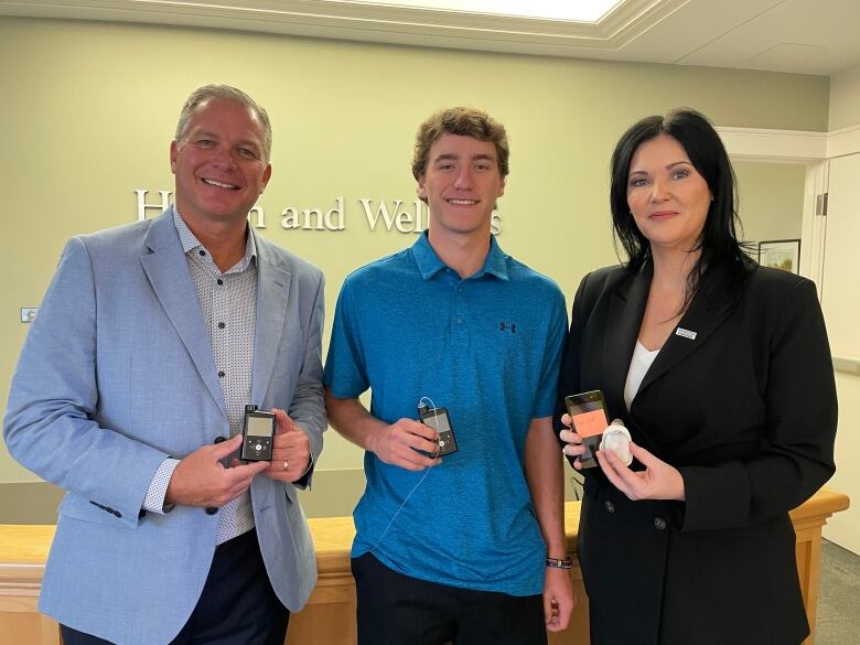 Three people stand holding insulin pumps and continuous glucose monitors. On the left, a man in a blue suit jacket and button up shirt, in the middle a young man with a blue polo shirt, and a woman with dark hair in a black pantsuit on the right.