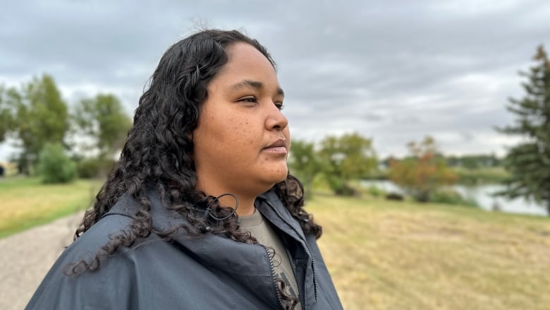 A woman in a black jacket with dark skin and curly black hair looks into the distance, with trees and a lake in the background. 