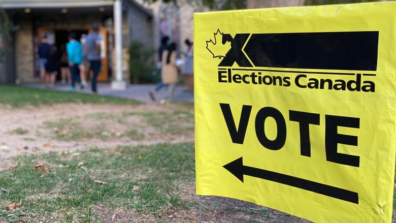 A yellow Elections Canada sign points to a local polling station. Voters line up in the background.