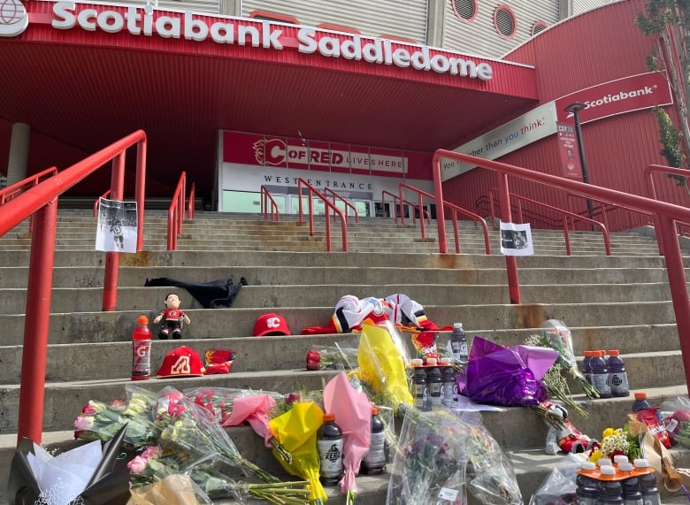 Flowers, Gatorade and various red Calgary hockey paraphernalia sit on grey steps. The sign for the scotiabank saddledome can be seen.