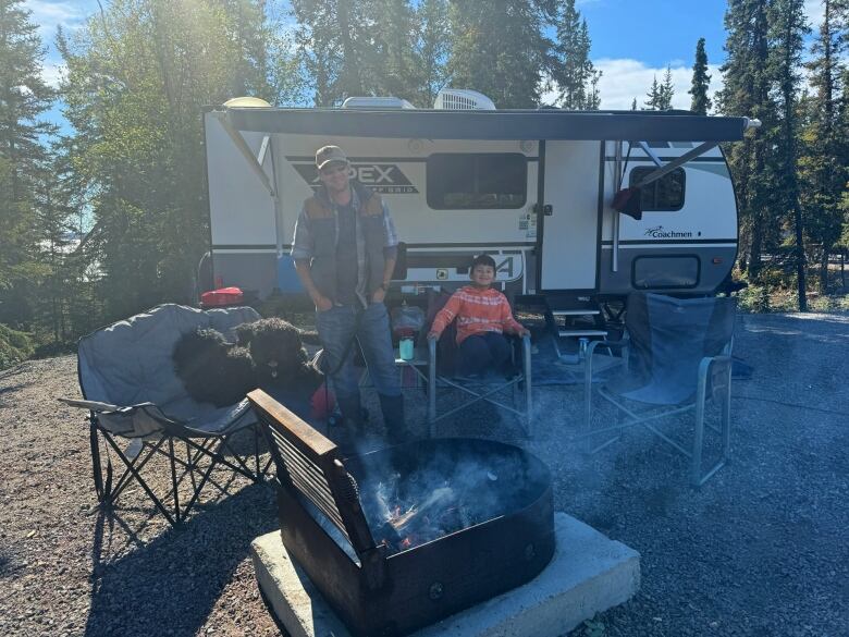 Man and child and dog sit on camping chairs in front of a camper