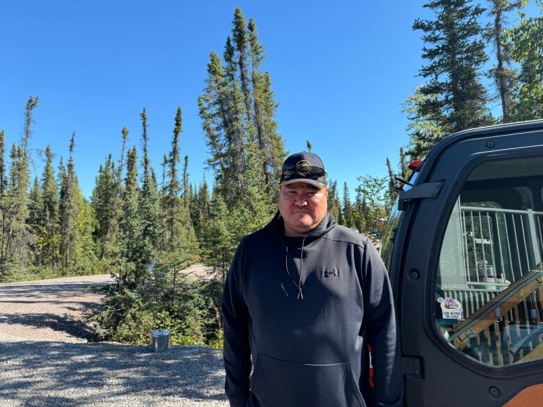 Brendan Camsell stands in front of trees on a sunny day. there is an ATV beside him