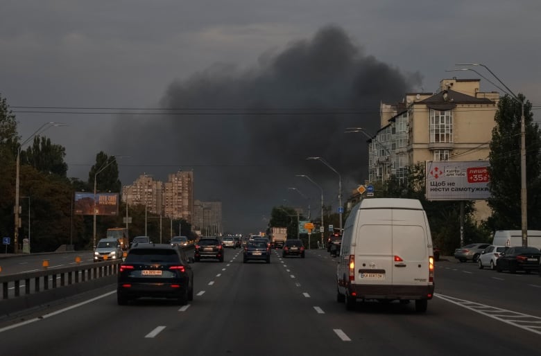 Smoke rises in the sky over a street with several cars.