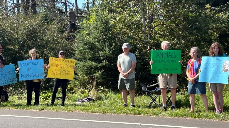 A group of people holding protest signs stand at the side of a road in a rural area.