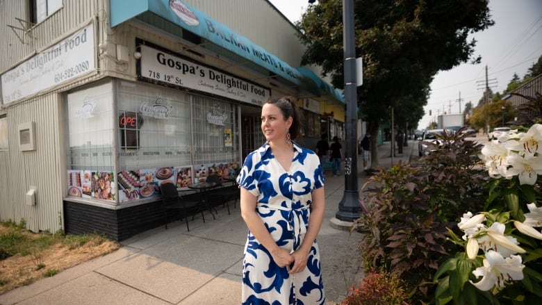 A woman in a blue and white dress stands infront of a Balkan deli. She's looking off to the side and smiling. 