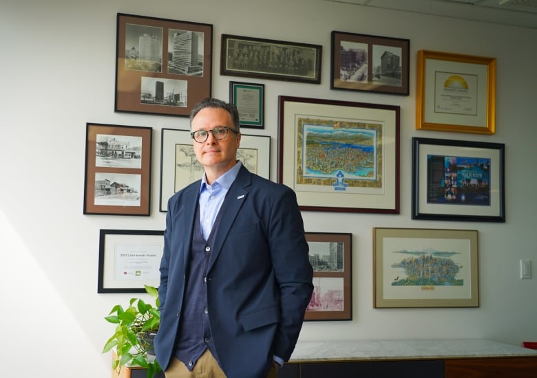 A man in a blue blazer stands infront of a wall with framed photos of buildings. 