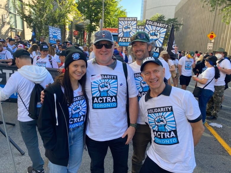 A group of actors in ACTRA t-shirts pose in front of a parade of people wearing similar shirts on a city street in daylight.