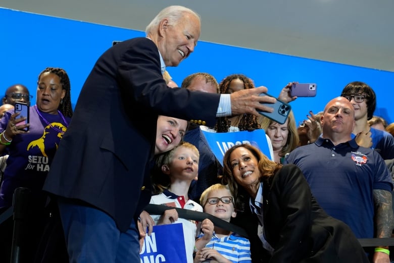 An older man in a suit jacket holds a phone as a woman and some small children pose for a photo.