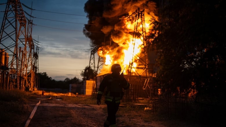 A large orange fire is shown near a hydro tower as a firefighter in uniform is shown in the foreground in shadow.