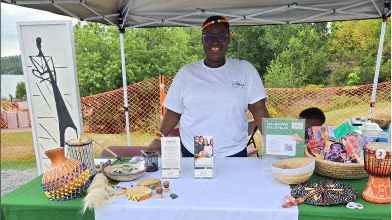 A Black woman standing behind an outdoor booth with various cultural objects.