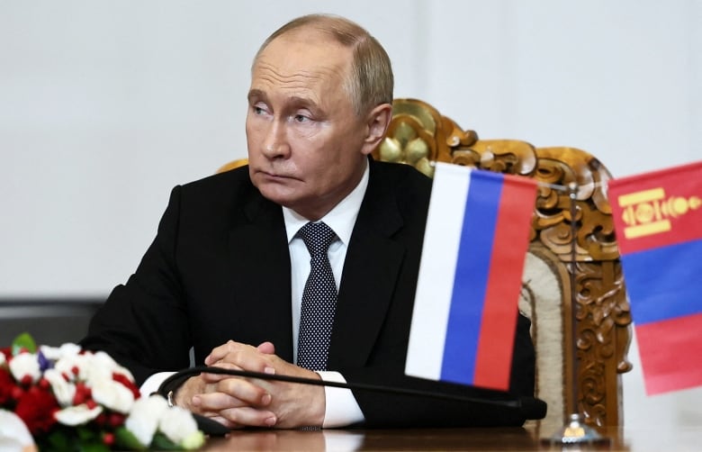 A stern man in a black suit with a black-and-white tie sits at a wooden table with his hands folded.