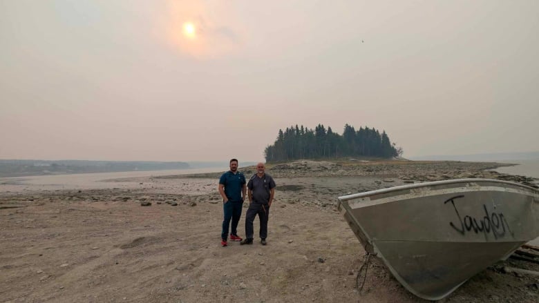 Two men stand on a sandy shoreline with a smoky background