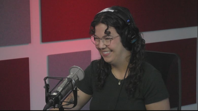 Woman with dark hair smiling, wearing black shirt and glasses. She's sitting at a desk in front of a microphone.