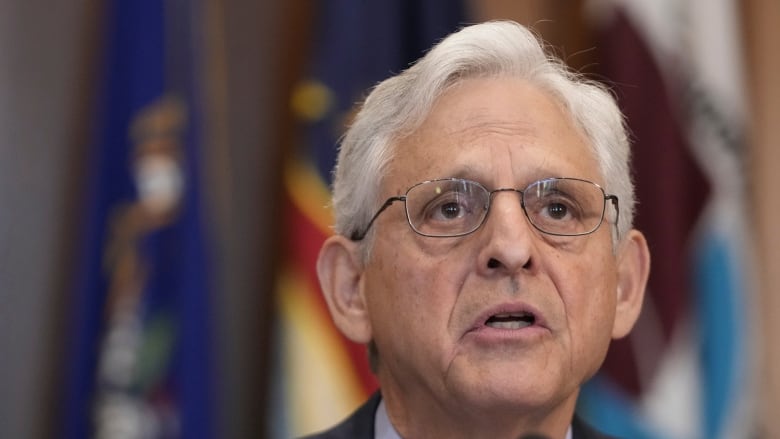 A man with grey hair, black glasses and a grey suit speaks into a microphone. 