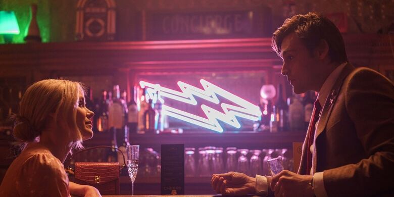 A man and a woman talk to one another at a bar with a neon light in the background.